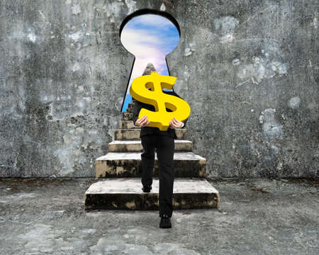 Man carrying golden dollar sign climbing old concrete stairs toward keyhole, with view of sky clouds.の写真素材