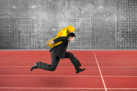 Businessman carrying 3D golden dollar sign running on red track, with city buildings doodles wall background.の写真素材