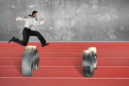 Man jumping over currency symbol obstacles on red running track, with gray concrete wall background.の写真素材