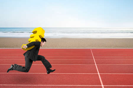 Businessman carrying 3D golden euro sign running on red track, with sky sea sand beach background.の写真素材