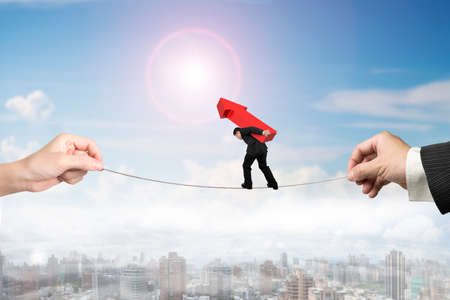 Businessman carrying red arrow sign, balancing on tightrope with man and woman hands holding two sides, on sun sky city landscape background.の写真素材