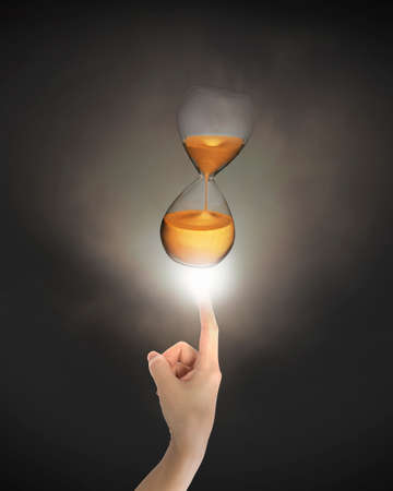 Human index finger pointing at hourglass with bright light, on black background.