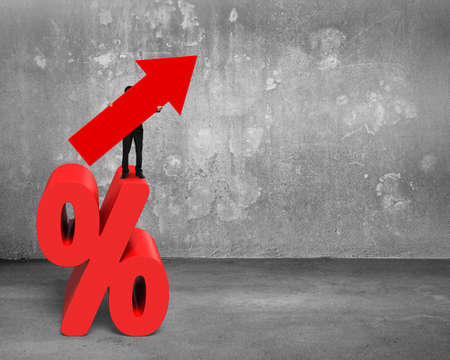Businessman holding red arrow symbol standing on percentage sign, on concrete wall and floor background.の写真素材