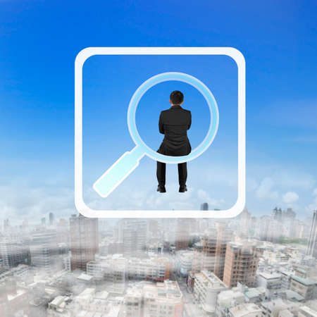 Rear view of businessman sitting on searching app icon, on blue sky cityscape background.