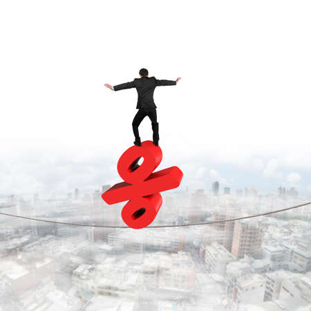 Businessman standing on red percentage sign balancing tightrope, with cloudy sky cityscape background.の写真素材
