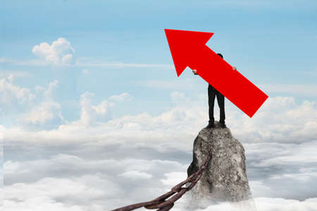 Man holding red arrow up sign, standing on mountain peak with rusty iron chain, on sky cloudscape...