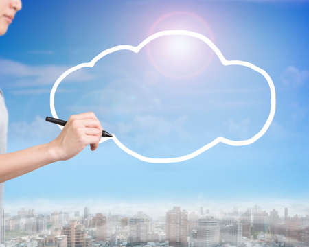 Woman hand holding pen drawing white cloud in sky, side view, with sunny cityscape background.の写真素材