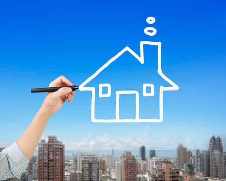Female hand holding pen drawing house shape clouds in sky, side view, with city buildings background.の写真素材