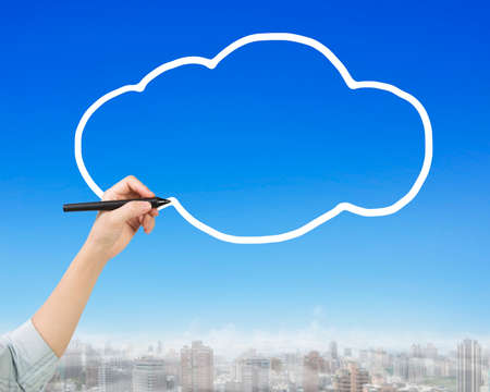 Female hand holding pen drawing white cloud in sky, side view, with city buildings background.の写真素材