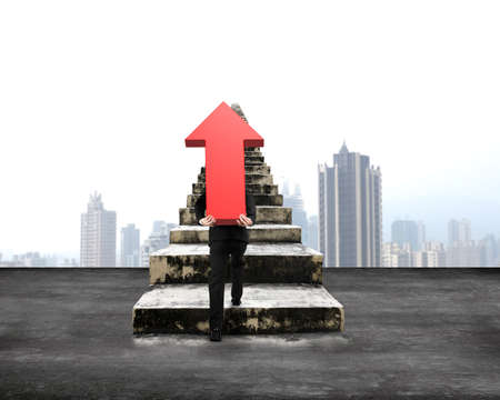 Man carrying red arrow up sign climbing old concrete stairs, with city skyscraper backgroundの写真素材