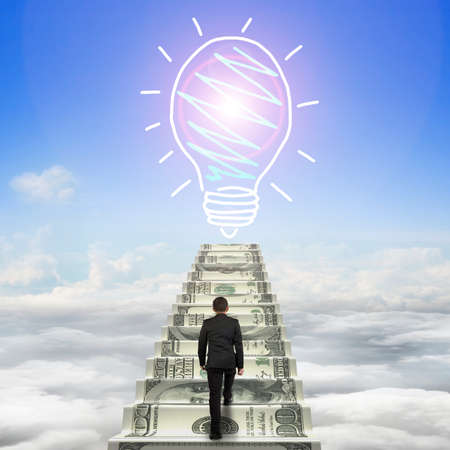 Businessman climbing the money stairs toward light bulb hand drawing, with sun sky cloudscape background.の写真素材