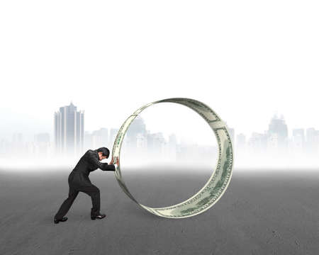 Businessman pushing a roll of dollar bills on concrete ground, with city skyscraper skyline background.の写真素材