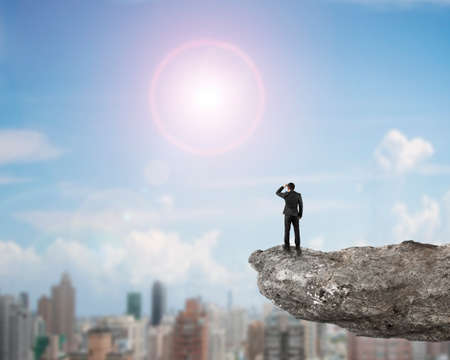 Rear view businessman standing gazing on cliff with sunny sky city buildings background.の写真素材