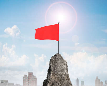 Red wavy flag on top of rocky mountain peak with sunlight and clouds in sky