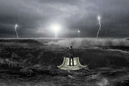 Man gazing on money boat floating in the ocean with storm, thunder, lightening and lighthouse in...