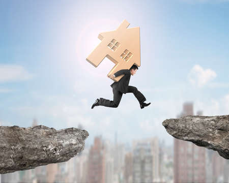 Man carrying wooden house jumping over two cliffs, with sunny sky city buildings background.の写真素材