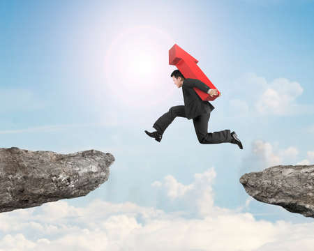 Man carrying red arrow sign jumping over two cliffs, on sunny sky clouds background.の写真素材