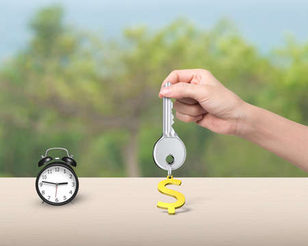 Woman hand holding silver key with golden dollar sign shape keyring, and alarm clock on the table.の写真素材