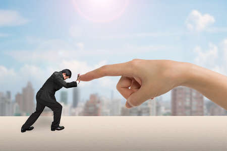Small man pushing against big woman hand forefinger on table, with city buildings background.の写真素材