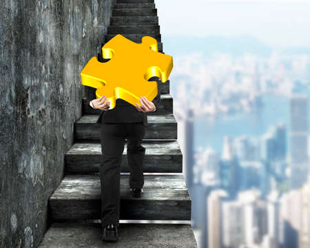 Businessman carrying big gold jigsaw puzzle piece and climbing on old dirty concrete stairs, with city buildings background.の写真素材