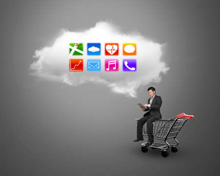 Man using digital tablet and sitting on the shopping cart with white cloud of colorful app icons, on gray background, on-line shopping concepts.の写真素材