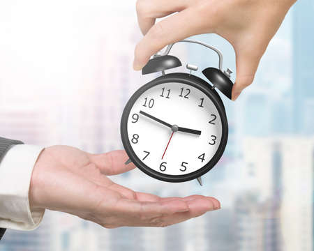 Woman hand giving one alarm clock to man hand, with city buildings background.