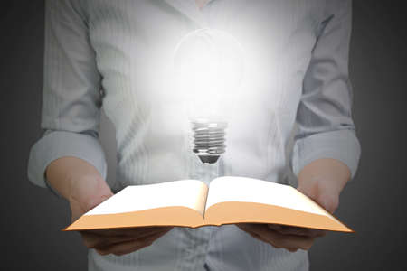 Woman hands holding the open book with bright light bulb illuminated the darkness, front view.