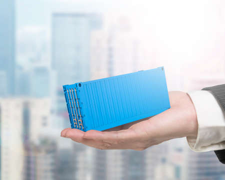 Human hand holding 3d blue cargo container, with sunny cityscape background.の写真素材
