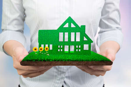 Green energy supply and solution concept, woman hands holding green house on grass land with mud and sunflowers, front view.の写真素材