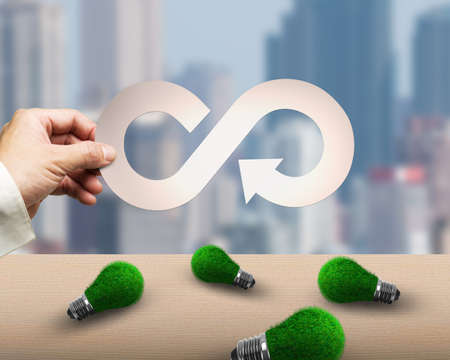 Circular economy and green energy concept. Man's hand holding metal arrow infinity recycling symbol, with green grass of light bulbs on the table and city buildings background.の写真素材