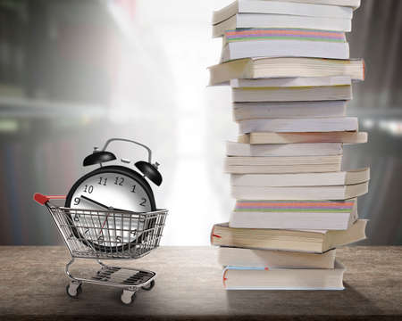 Alarm clock in shopping cart with stack of books on wooden desk and blur bookshelf background. Concept of buying time for reading many books.の写真素材