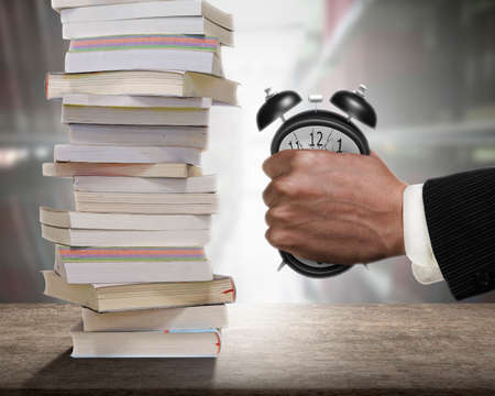 Concept of many books need to read, but not enough time. Man fist clenched the deformed alarm clock in anger with stack of books on wooden desk and bookshelf background.の写真素材