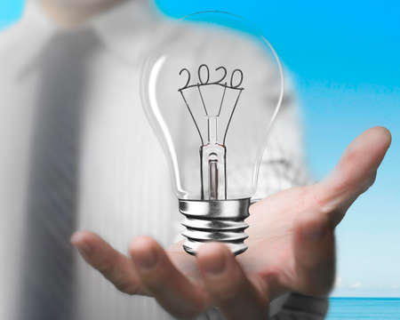 Businessman hand holding light bulb with 2020 wire shape, front view, blue sky sea background.の写真素材