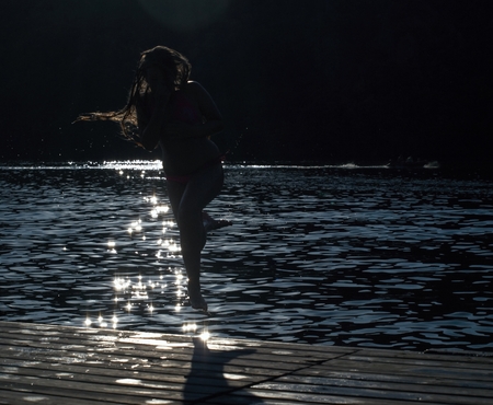 The girl is jumping into the water from the wooden pier in backlight from reflections on the waterの写真素材