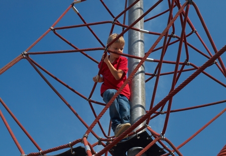 Blond little boy in red T-shirt on pyramid net climber under blue sky.の写真素材