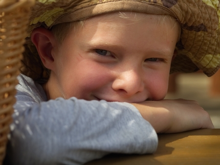 Close up of smiling little boy in an outdoor hat rests on a wooden table where a wicker basket is laid.の写真素材