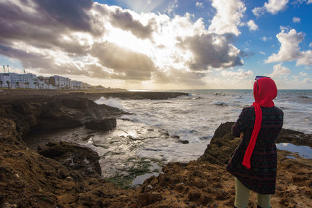 North Africa. Morocco. Rabat. A young woman wearing a red veil watches the rough sea on the coast in Rabatのeditorial素材