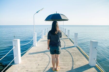 Teenage woman standing holding an black umbrella on white cement bridge fishing pier extending into the sea at Khlong Ta Kok Bang Pu Samut Prakan, Thailand.の写真素材