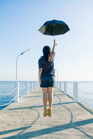 Teenage woman jumping holding an black umbrella on white cement bridge fishing pier extending into the sea at Khlong Ta Kok Bang Pu Samut Prakan, Thailand.の写真素材