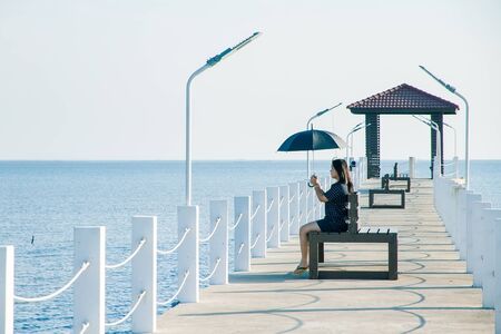 Teenage woman sitting holding an black umbrella on white cement bridge fishing pier extending into the sea at Khlong Ta Kok Bang Pu Samut Prakan, Thailand.の写真素材