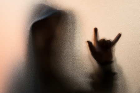 Shadow blur of horror man in jacket with hood.Finger do as a symbol love on the glass.Dangerous man behind the frosted glass.Mystery man.Halloween background.Blur pictureの写真素材