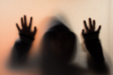 Shadow blur of horror man in jacket with hood.Hands on the glass.Dangerous man behind the frosted glass.Mystery man.Halloween background.Blur picture.の写真素材