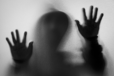 Shadow blur of horror man in jacket with hood.Hands on the glass.Dangerous man behind the frosted glass.Mystery man.Halloween background.Black and white picture.Blur pictureの写真素材