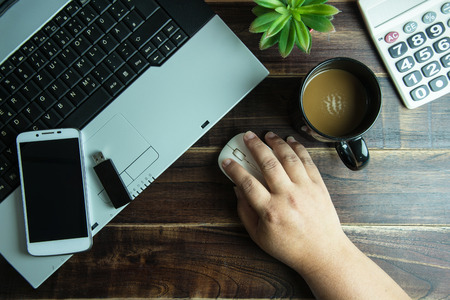 Top view of office stuff hand on wireless mouse with smart phone laptop and coffee cup on wooden table.Concept workplaceの写真素材