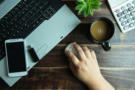 Top view of office stuff hand on wireless mouse with smart phone laptop and coffee cup on wooden table.Concept workplaceの写真素材