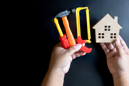 Man's hand holding wooden house toy and construction tools toy on black background with copy space.Real estate concept, New house concept, Finance loan business conceptの写真素材