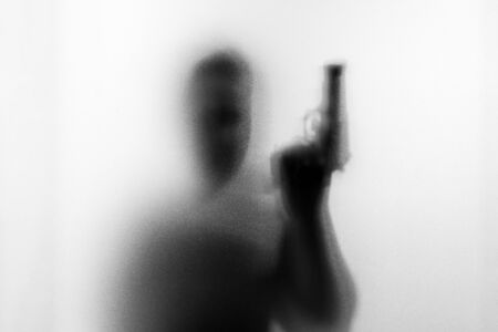 Shadow of horror man killer with a gun in his hand.Dangerous man behind the frosted glass.Mystery man.Halloween background.Black and white picture.Blur pictureの写真素材
