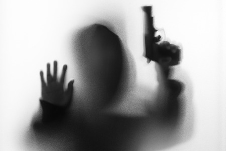 Shadow blur of horror man in jacket with hood gun in his hand.Dangerous man behind the frosted glass.Mystery man.Halloween background.Black and white picture.Blur pictureの写真素材