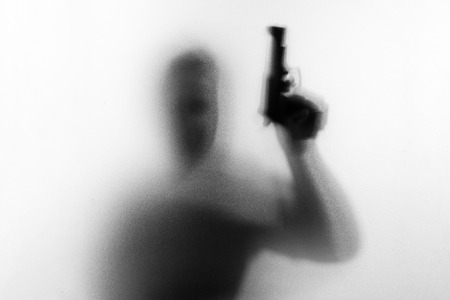 Shadow of horror man killer with a gun in his hand.Dangerous man behind the frosted glass.Mystery man.Halloween background.Black and white picture.Blur pictureの写真素材