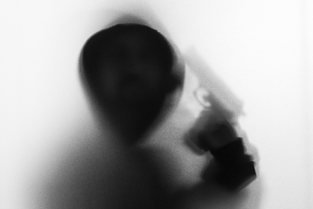 Shadow blur of horror man in jacket with hood gun in his hand.Dangerous man behind the frosted glass.Mystery man.Halloween background.Black and white picture.Blur pictureの写真素材